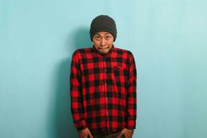 Embarrassed young Asian man bites his lower lip while looking at camera, isolated on blue background photo