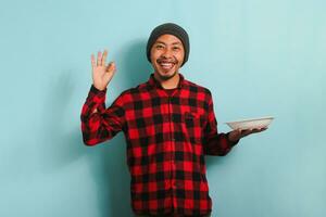 contento joven asiático hombre con un gorro sombrero y un rojo tartán franela camisa es sonriente y diciendo Hola mientras participación un vacío blanco lámina, aislado en un azul antecedentes foto