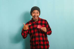 Excited young Asian man with a beanie hat and red plaid flannel shirt is pointing to his bank credit cards in his hands while looking at camera and standing against a blue background photo