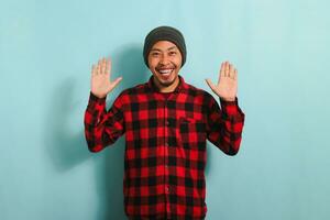 An attractive and friendly young Asian man with a beanie hat and a red plaid flannel shirt smiles, waves, and says hello, hi, or bye while standing against a blue background photo