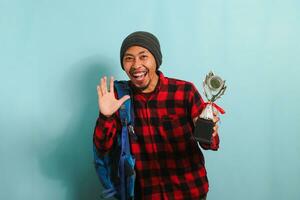 Excited Young Asian man student wearing a backpack, beanie hat, and red plaid flannel shirt, holding trophy, smiling at the camera, isolated on a blue background photo