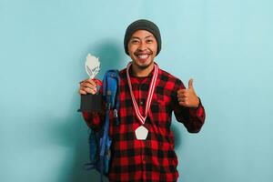 Excited young Asian man student wearing backpack, beanie hat, and red plaid flannel shirt, giving thumbs up while holding a trophy and rejoicing in success and achievement, isolated on blue background photo