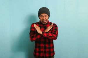 An angry young Asian man with a beanie hat and a red plaid flannel shirt stands with his arms crossed shouting, displaying a refusal, denial, and stop gesture while standing against a blue background. photo