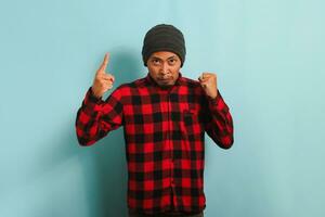 An angry young Asian man with a beanie hat and a red plaid flannel shirt is pointing, shouting, and yelling at the camera. He is isolated on a blue background photo