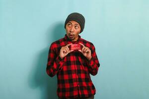 Young Asian man with a beanie hat and red plaid flannel shirt is holding bank credit cards in his hands, with a surprised expression looking at copy space while standing against a blue background. photo