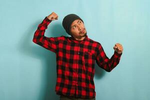 Young Asian man with beanie hat and red plaid flannel shirt making strong gesture, showing strength by flexing his arms and muscles, looking aside at empty copy space, isolated on a blue background photo