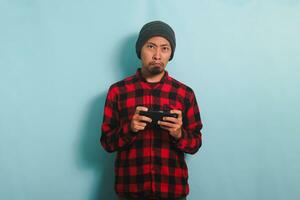 Bored young Asian man with a beanie hat and a red plaid flannel shirt is playing a boring game and watching an uninteresting video online on his smartphone, isolated on a blue background photo