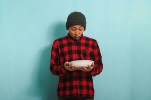 sorprendido joven asiático hombre con un gorro sombrero y un rojo tartán franela camisa es participación un vacío blanco plato en su mano, aislado en un azul antecedentes foto