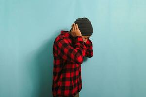 Young Asian man with beanie hat and red plaid flannel shirt holds his hand near his ear, suffering from tinnitus, a throbbing earache, and fatigue due to noise, while standing against blue background photo