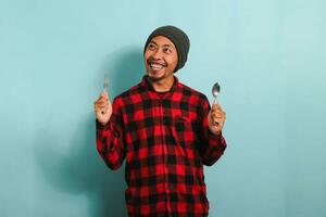 Young Asian man with beanie hat and red plaid flannel shirt holding a spoon and fork, looking happy and eager to eat, isolated on a blue background photo