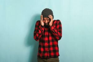 An embarrassed young Asian man with a beanie hat and a red plaid flannel shirt feels bad, guilty, and sorry while standing against a blue background photo