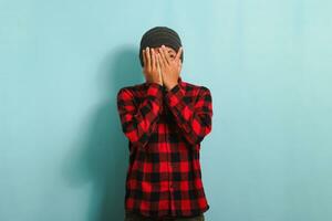 An ashamed and embarrassed young Asian man with a beanie hat and a red plaid flannel shirt facepalms, covering his face with his hand while standing against a blue background photo