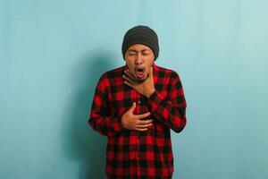 Ill Young Asian man with a beanie hat and red plaid flannel shirt is coughing and sneezing, suffering from a cough, flu, and allergies, while standing against a blue background photo