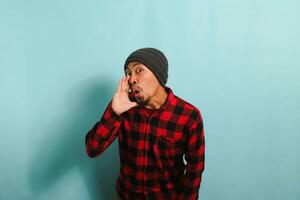 A young Asian man with a beanie hat and a red plaid flannel shirt keeps his hand on his mouth, telling a secret and spreading a rumor through whispering while standing against a blue background photo