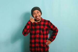 An excited young Asian man wearing a beanie hat and a red plaid flannel shirt makes a CALL ME gesture with his hand, isolated on a blue background photo