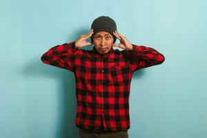 An unhappy young Asian man with a beanie hat and a red plaid flannel shirt grabs his head, staring alarmed at the camera, and feeling anxious while standing against a blue background photo