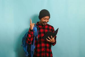 Excited young Asian student with a beanie hat and a red plaid flannel shirt, wearing a backpack, is pointing his finger up, feeling happy as he gets a good idea while standing against blue background photo