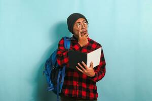 A thoughtful young Asian student man with beanie and red plaid flannel shirt, wearing a backpack and glasses, is thinking while holding books looking for ideas while standing against a blue background photo