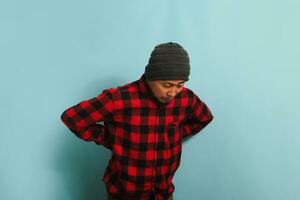 A young Asian man with a beanie hat and a red plaid flannel shirt is suffering from a backache, experiencing back pain. He is isolated on a blue background photo