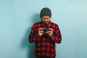 Confused young Asian man with a beanie hat and a red plaid flannel shirt feels disappointed and annoyed after losing while playing an online game on his smartphone, isolated on a blue background photo