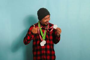 A young Asian man with a medal, a beanie hat, and a red plaid flannel shirt points a finger to himself, smiling with happiness and pride while standing against a blue background photo