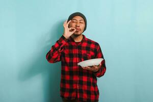 satisfecho joven asiático hombre con gorro sombrero y rojo tartán franela camisa es haciendo delicioso mano gesto a Rápido cómo bueno el comida es mientras participación un vacío blanco lámina, aislado en un azul antecedentes foto