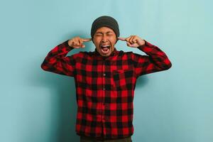 Angry young Asian man with a beanie hat and a red plaid flannel shirt covers his ears, feeling the painful ache of an earache and suffering from otitis due to loud noise, isolated on blue background photo