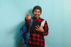A smiling young Asian student with a beanie hat and a red plaid flannel shirt, wearing a backpack, is pointing at the camera while standing against a blue background photo