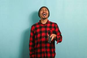 Happy young Asian man with a beanie hat and red plaid flannel shirt bursting out laughing while watching TV, isolated on a blue background photo