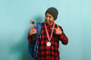 Excited young Asian man student wearing a backpack, beanie hat, and red plaid flannel shirt, pointing to the trophy in his hand while rejoicing in success and achievement, isolated on blue background photo