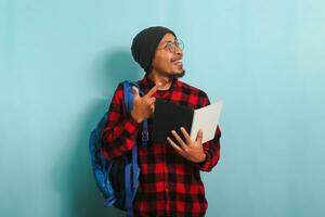 Excited young Asian student with a beanie hat and a red plaid flannel shirt, wearing a backpack and glasses, is holding books and pointing aside the copy space while standing against a blue background photo
