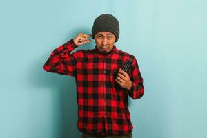 Scared young Asian man with a beanie hat and red plaid flannel shirt covering his ears feeling nervous while watching a thriller movie, isolated on a blue background photo