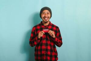 Excited young Asian man with a beanie hat and red plaid flannel shirt is holding bank credit cards in his hands while standing against a blue background. Finance, loan, savings concept photo