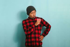 Young Asian man with a beanie hat and red plaid flannel shirt is holding bank credit cards in his hands, with a surprised expression on his face while standing against a blue background. photo
