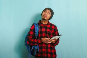 A pensive young Asian student with a beanie hat and a red plaid flannel shirt, wearing a backpack, is deep in thought, looking up at the copy space while standing against a blue background photo