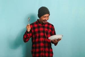 emocionado hambriento joven asiático hombre con gorro sombrero y rojo tartán franela camisa participación un cuchara, mirando a un vacío blanco plato en su mano, aislado en un azul antecedentes foto