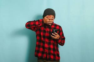 gracioso joven asiático hombre con gorro sombrero y rojo tartán camisa es echar un vistazo a inapropiado contenido en su teléfono, conmocionado y cubierta su rostro, mirando mediante su dedos, aislado en azul fondo foto