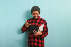 A smiling young Asian man with a beanie hat and a red plaid flannel shirt is pointing to a book in his hand while standing against a blue background photo