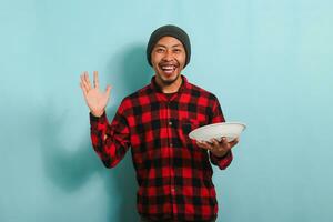 contento joven asiático hombre con un gorro sombrero y un rojo tartán franela camisa es sonriente y diciendo Hola mientras participación un vacío blanco lámina, aislado en un azul antecedentes foto