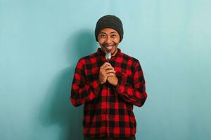 Happy Young Asian man with beanie hat and red plaid flannel shirt holding a spoon near his mouth, feeling excited and eager to eat, isolated on a blue background photo