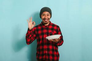 Satisfied Young Asian man with beanie hat and red plaid flannel shirt is making delicious hand gesture to express how good the food is while holding an empty white plate, isolated on a blue background photo