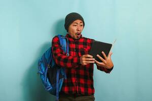 An excited young Asian student with a beanie hat and a red plaid flannel shirt, wearing a backpack, is pleasantly surprised by a book he read while standing against a blue background photo