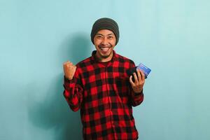 emocionado joven asiático hombre con gorro sombrero y rojo tartán franela camisa participación un billetera y crédito tarjeta, aislado en un azul antecedentes foto