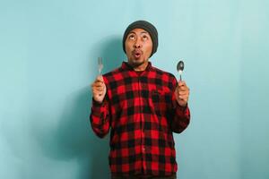 Excited Young Asian man with beanie hat and red plaid flannel shirt looking up with a surprised expression while holding spoon and fork, isolated on blue background photo