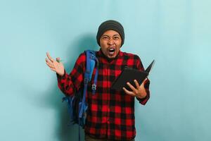 Angry young Asian student with a beanie hat and a red plaid flannel shirt, wearing a backpack, looks shocked after realizing he forgot to study before the exam while standing against a blue background photo