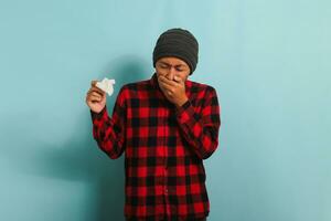 Young Asian man with a beanie hat and red plaid flannel shirt is coughing and sneezing, suffering from a cough, flu, and allergies, while holding a tissue, isolated on blue background photo