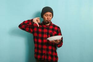 disgustado joven asiático hombre con un gorro sombrero y un rojo tartán franela camisa es dando un pulgares abajo, dando el comida un malo revisar, mientras participación un vacío blanco lámina, aislado en un azul antecedentes foto