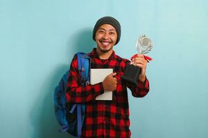 Happy young Asian man student wearing a backpack, beanie hat, and red plaid flannel shirt, holding a silver trophy, rejoicing in his success and achievement, isolated on a blue background. photo