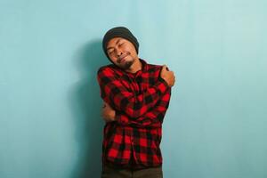 joven asiático hombre con gorro sombrero y rojo tartán franela camisa abrazos él mismo, expresando felicidad y positividad, mientras en pie en contra un azul antecedentes. yo amor y yo cuidado concepto. foto