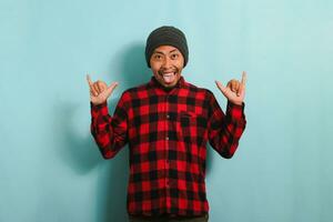 Happy young Asian man with a beanie hat and red plaid flannel shirt showing the rock and roll gesture with his fingers and sticking out his tongue, isolated on a blue background photo